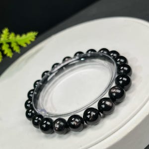 Puede incluir: Una pulsera de cuentas negras con un acabado brillante. La pulsera está hecha de cuentas redondas y pulidas. Las cuentas están ensartadas en una banda elástica transparente. La pulsera se exhibe sobre una superficie blanca.
