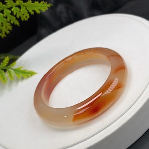 May include: A smooth, polished bangle bracelet crafted from translucent agate stone. The bracelet features a rounded profile with a gradient of warm colors, transitioning from soft beige and cream tones to deep, vibrant streaks of reddish-orange.