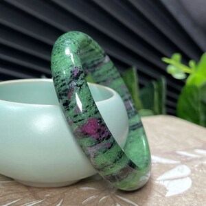 Puede incluir: Un brazalete de piedra verde, negro y rosa. El brazalete est&aacute; hecho de piedra pulida y tiene una superficie lisa y redondeada.