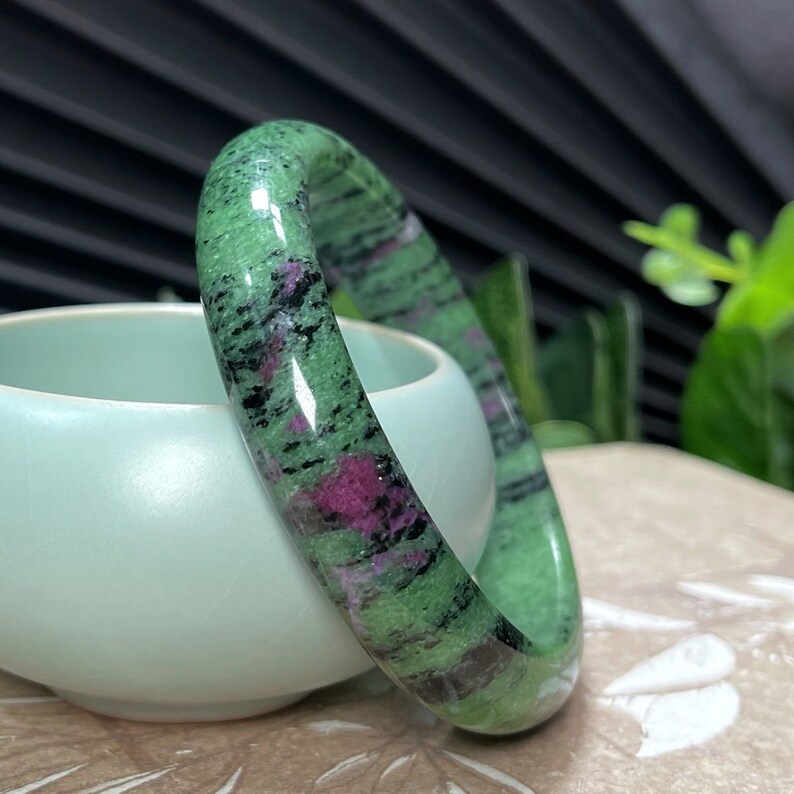 Puede incluir: Un brazalete de piedra verde, negro y rosa. El brazalete est&aacute; hecho de piedra pulida y tiene una superficie lisa y redondeada.