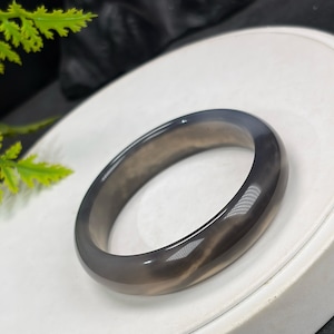 May include: A polished, gray gemstone bangle bracelet. The smooth, circular bracelet has a marbled appearance with shades of gray and brown. The jewelry is displayed on a white surface, showcasing its elegant design. This accessory is suitable for everyday wear or special occasions.
