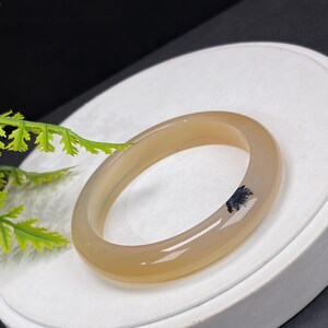 Könnte beinhalten: Ein beigefarbenes Armreif-Armband aus Edelstein mit glatter Oberfläche. Das kreisförmige Armband hat einen dunkelblauen Einschluss. Es liegt auf einer weißen Oberfläche.