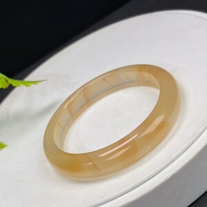 May include: A translucent, light brown bangle bracelet. The smooth, polished surface of the bracelet reflects light, showcasing its natural variations. The bracelet is a simple, elegant piece of jewelry.