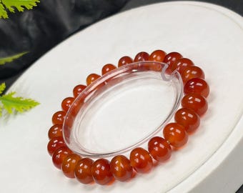 May include: A bracelet with polished, reddish-orange beads. The beads are strung together, forming a circular shape. The bracelet is displayed on a white surface, with a clear plastic support structure holding the bracelet in place.