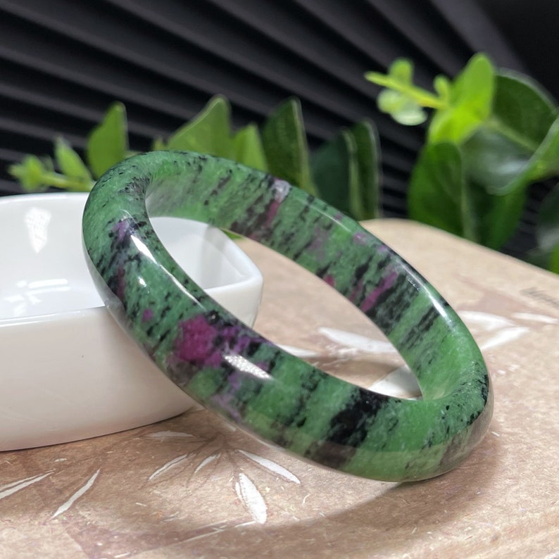 Puede incluir: Un brazalete de jade verde, negro y rosa. El brazalete est&aacute; hecho de piedra pulida y tiene una superficie lisa y redondeada.