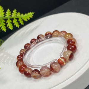 Op de afbeelding: Een kralenarmband met gepolijste, ronde edelstenen in tinten rood, bruin en crème. De armband is geregen op een transparant elastisch koord en bevat een goudkleurige spacer met heldere kristallen.