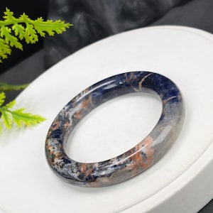 May include: A polished bangle bracelet crafted from natural stone, featuring a smooth, rounded surface with a marbled pattern of deep navy blue, translucent grey, and streaks of burnt orange throughout the entire circular band.