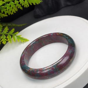 May include: A polished, circular bangle bracelet crafted from a smooth, solid stone featuring a mottled pattern of deep magenta, dark red, and teal blue hues. The surface of the bangle has a glossy, reflective finish with a rounded, thick profile.