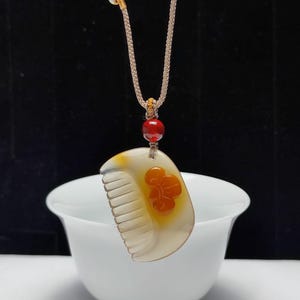 Può includere: Una collana con pendente a forma di pettine. Il ciondolo bianco sporco presenta dettagli floreali arancioni e un design a pettine. Una perla rossa è sopra il ciondolo, attaccata a un cordino beige. Lo sfondo è scuro e la collana è esposta in una ciotola bianca.
