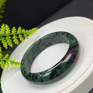 May include: A polished gemstone bangle. The bracelet is a mix of green and red hues, with a smooth, rounded shape. The bracelet is displayed on a white surface. The bracelet's diameter is approximately 6.5 cm.
