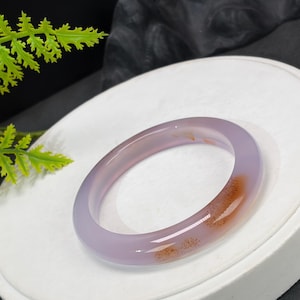 Peut inclure: Un bracelet jonc lisse et translucide de couleur lavande, fabriqué en pierre polie. L'anneau circulaire présente une inclusion ou une tache de couleur brun rougeâtre distincte et naturelle sur un côté, contrastant avec la teinte violet clair.
