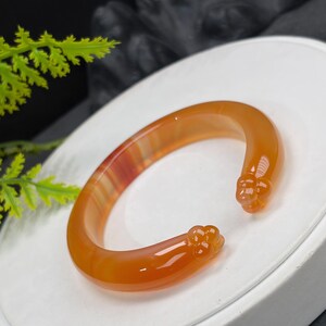 Könnte beinhalten: Orangenes Achat-Armband mit einem geschnitzten Blumendesign an jedem Ende. Das Armband hat eine warme, durchscheinende Farbe mit roten und weißen Bändern. Das offene Manschettendesign ermöglicht ein einfaches Tragen. Ein einzigartiges Schmuckstück.