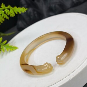 Könnte beinhalten: Ein hellbraunes offenes Armreif-Armband mit poliertem Finish. Das Armband hat ein glattes, geschwungenes Design mit einer leichten Öffnung. Die Farbe geht von hell- zu dunkelbraun über. Das Armband besteht aus einem durchscheinenden Material.