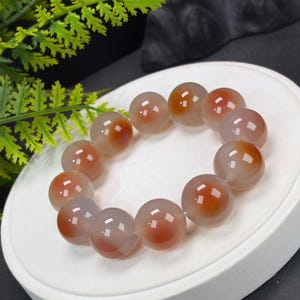 Könnte beinhalten: Ein Perlenarmband aus glatten, durchscheinenden, kugelförmigen Perlen. Jede Perle weist einen deutlichen, zentrierten rötlich-orangen Einschluss in einer blassen, halbtransparenten grauen Basis auf. Die Perlen sind kreisförmig angeordnet.