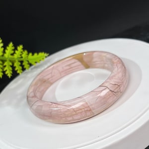 以下が含まれることがあります： ピンクとブラウンのパターンが施された、研磨された石製のバングルブレスレットです。