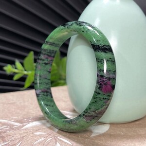 Puede incluir: Un brazalete de piedra verde, negro y rosa. El brazalete est&aacute; hecho de piedra pulida y tiene una superficie lisa y redondeada.