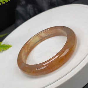 May include: A translucent, oval-shaped bracelet in shades of amber and cream. The polished surface of the stone reflects light, showcasing the natural patterns within. This piece of jewelry is a classic accessory.