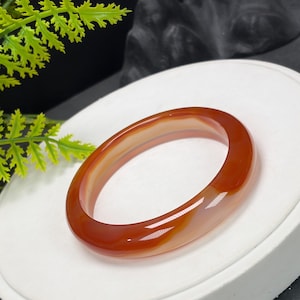 May include: A smooth, polished bangle bracelet crafted from translucent carnelian agate. The circular jewelry piece features a rich, deep orange-red color with natural, creamy white banding patterns visible throughout the stone's polished surface.