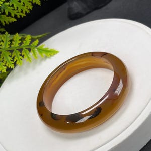 以下が含まれることがあります： 半透明の茶色いメノウ石で作られた、滑らかで磨き上げられたバングルブレスレット。円形のバンドには、半透明の素材の中に自然な濃い茶色と黒のインクルージョンが含まれており、全体にユニークで有機的な模様を作り出しています。