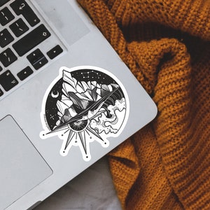 May include: Black and white sticker featuring a mountain range, a crescent moon, a sun with an eye, and waves. The sticker is on a silver laptop.