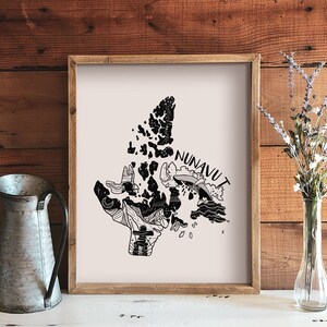 K&ouml;nnte beinhalten: Ein gerahmter Kunstdruck mit einer Schwarz-Wei&szlig;-Illustration der Region Nunavut in Kanada. Das Kunstwerk enth&auml;lt das Wort "NUNAVUT" und verschiedene grafische Elemente. Eine Metallkanne und eine Vase mit Blumen befinden sich im Vordergrund.