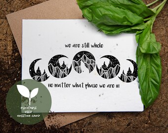 May include: A white greeting card with the text "we are still whole, no matter what phase we are in." The card features a black and white design of moon phases and mountains. A green plant is in the background.
