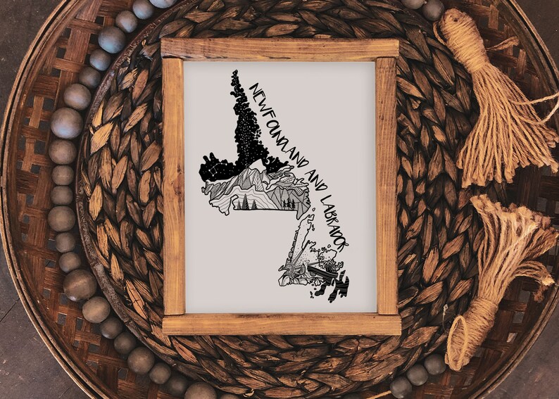 May include: A framed print with a black and white illustration of the Newfoundland and Labrador map. The artwork includes the text "NEWFOUNDLAND AND LABRADOR". The frame is made of wood. The print is displayed on a woven surface.