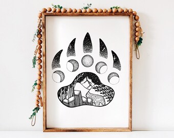 May include: Framed black and white art print depicting a bear paw print. The paw print design includes a mountain landscape and moon phases. The wooden frame is decorated with wooden beads and greenery.