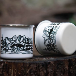 May include: Two white enamel mugs with a silver rim, featuring a black and white mountain and forest design. One mug is upright, the other is on its side. The mugs are on a wooden surface.