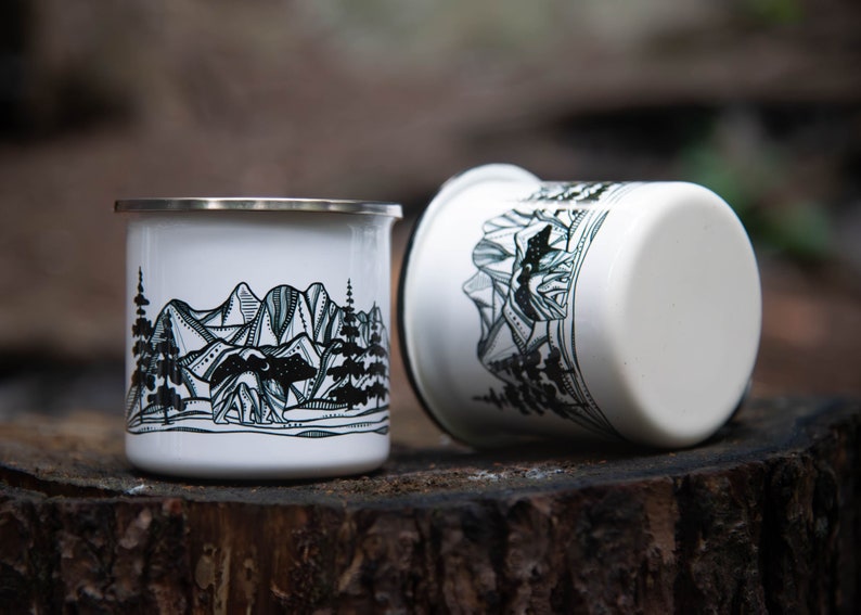 May include: Two white enamel mugs with a silver rim, featuring a black and white mountain and forest design. One mug is upright, the other is on its side. The mugs are on a wooden surface.