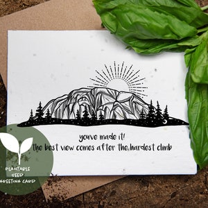 May include: A black and white plantable seed greeting card with a mountain illustration and the text "you've made it! the best view comes after the hardest climb." The card is on a brown surface with green leaves.