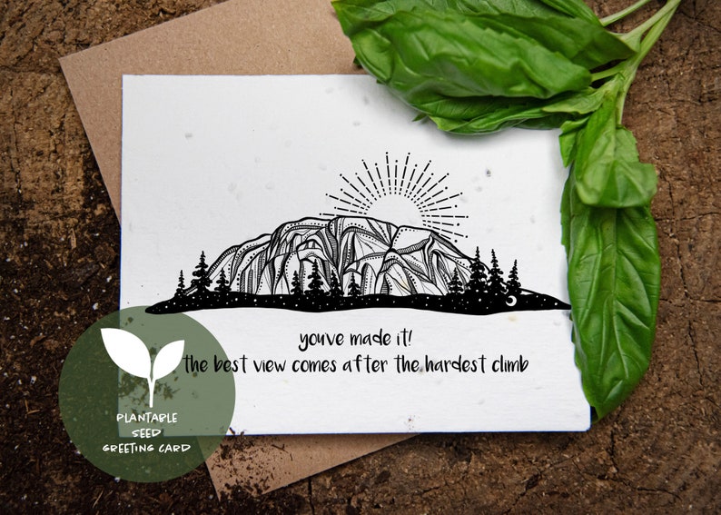 May include: A black and white plantable seed greeting card with a mountain illustration and the text "you've made it! the best view comes after the hardest climb." The card is on a brown surface with green leaves.