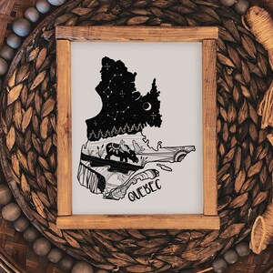 May include: A framed art print featuring the silhouette of the Quebec province, with a night sky design and moose illustration. The word "QUEBEC" is written at the bottom. The frame is made of wood and is set on a woven basket.