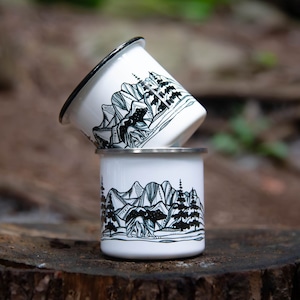 May include: Two white enamel camping mugs stacked on a wooden surface. Each mug features a black and white mountain and bear illustration. The rims are black and silver.