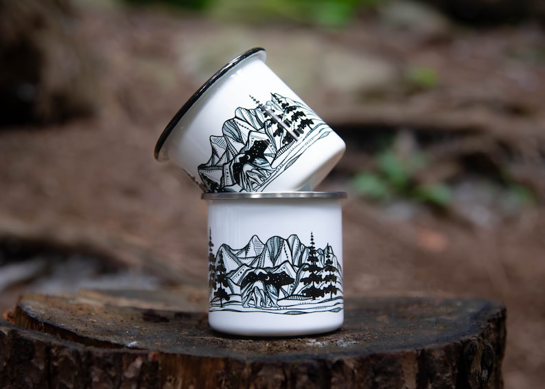 May include: Two white enamel camping mugs stacked on a wooden surface. Each mug features a black and white mountain and bear illustration. The rims are black and silver.