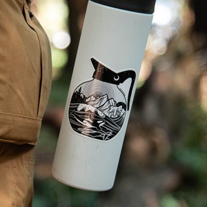 May include: A white stainless steel water bottle with a black and white sticker of a coffee pot with a mountain scene. The sticker has a crescent moon and stars.