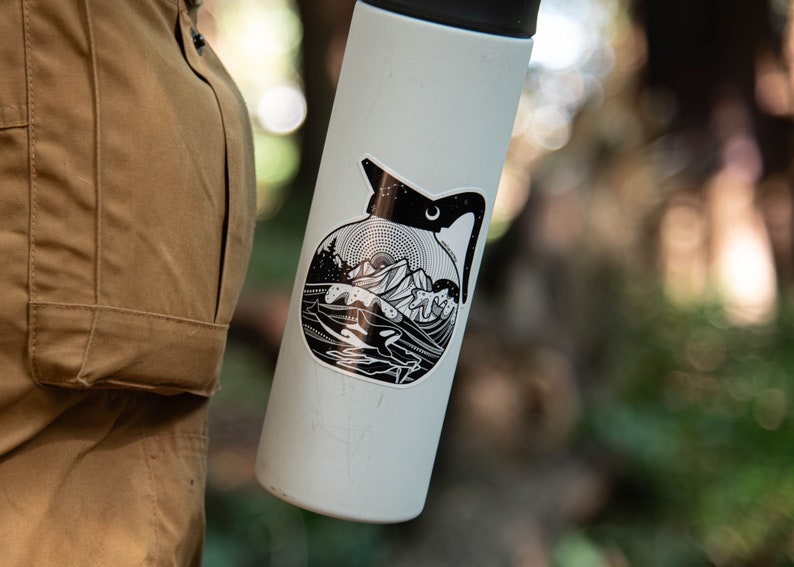 May include: A white stainless steel water bottle with a black and white sticker of a coffee pot with a mountain scene. The sticker has a crescent moon and stars.