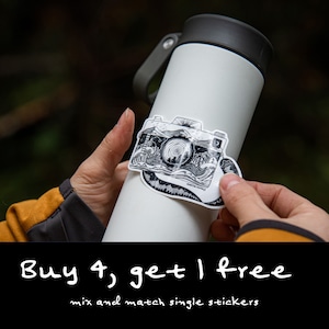 K&ouml;nnte beinhalten: Eine wei&szlig;e Wasserflasche mit schwarzem Deckel und Griff. Eine Person bringt einen schwarz-wei&szlig;en Aufkleber mit Kameradesign an. Der Text "Buy 4, get 1 free" befindet sich unten.