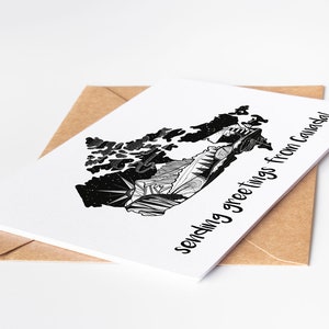 May include: A white greeting card with a black and white illustrated map of Canada, with the text "sending greetings from Canada!" The card is placed on a brown envelope, set against a white background.