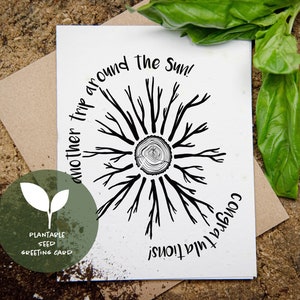 Può includere: Un biglietto di auguri bianco con un disegno ad anello di albero nero e le parole "another trip around the Sun!" e "Congratulations!". Un cerchio verde con una grafica a foglia e le parole "Plantable Seed Greeting Card" si trova in basso a sinistra. Il biglietto è su una superficie di legno.
