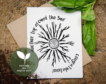 May include: A white greeting card with a black tree ring design and the words "another trip around the Sun!" and "Congratulations!" A green circle with a leaf graphic and the words "Plantable Seed Greeting Card" is in the lower left corner. The card is on a wooden surface.