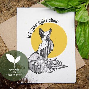 May include: A white greeting card featuring a black and white fox illustration, a yellow sun, and the words "let your light shine." The card is a plantable seed greeting card. The background is a brown wooden surface.