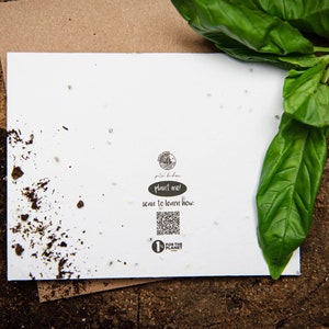 May include: A white card with the text "Plant me!" and a QR code, resting on a brown surface. Green basil leaves are in the upper right corner. The card has small seeds embedded in it.