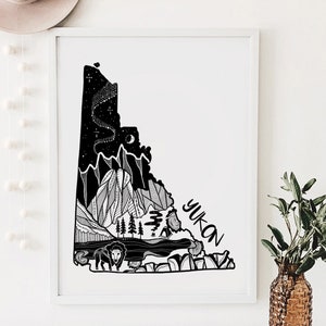 May include: Black and white art print of the Yukon territory. The artwork features a detailed illustration of mountains, trees, a bison, and the text "YUKON". The night sky is depicted with stars and a crescent moon. The print is framed in a white border.