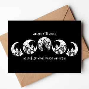 May include: Black greeting card with a white illustration of moon phases and mountains. The text reads "we are still whole no matter what phase we are in". The card is inside a brown envelope.