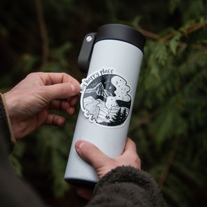 May include: A white stainless steel water bottle with a black lid and a black and white sticker that says "My happy place" with a mountain and forest scene.