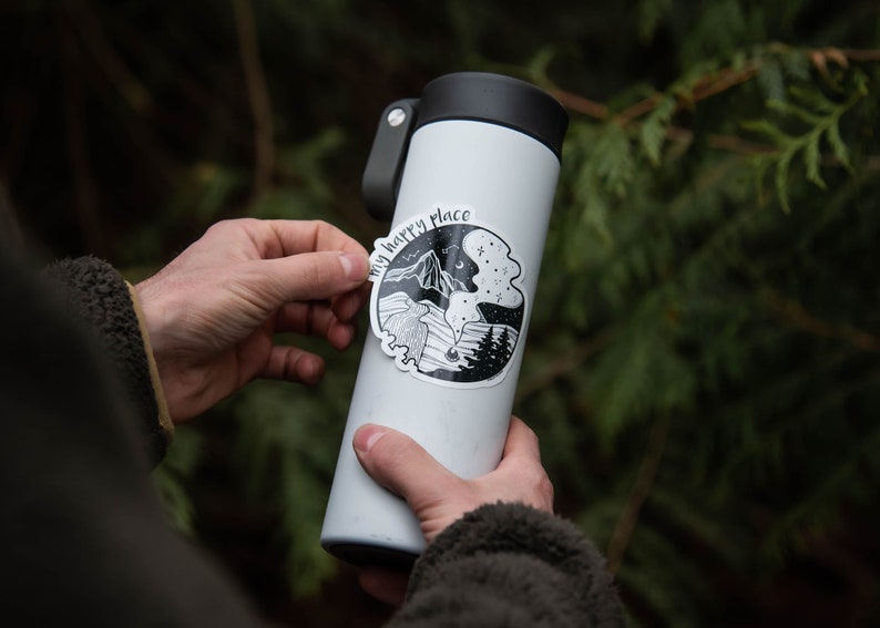 May include: A white stainless steel water bottle with a black lid and a black and white sticker that says "My happy place" with a mountain and forest scene.