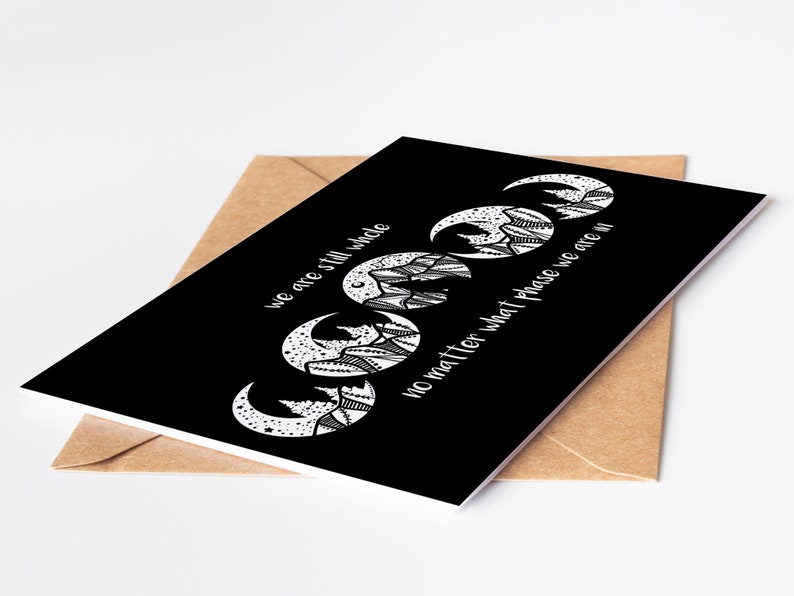 May include: A black greeting card with a white crescent moon design and the text "we are still whole no matter what phase we are in." The card is inside a brown envelope.