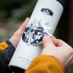 May include: A white stainless steel water bottle with a black and white sticker of a mountain scene with a sun and waves. The bottle has the text "MICRO" and "24 oz / 720 ml" printed on it.