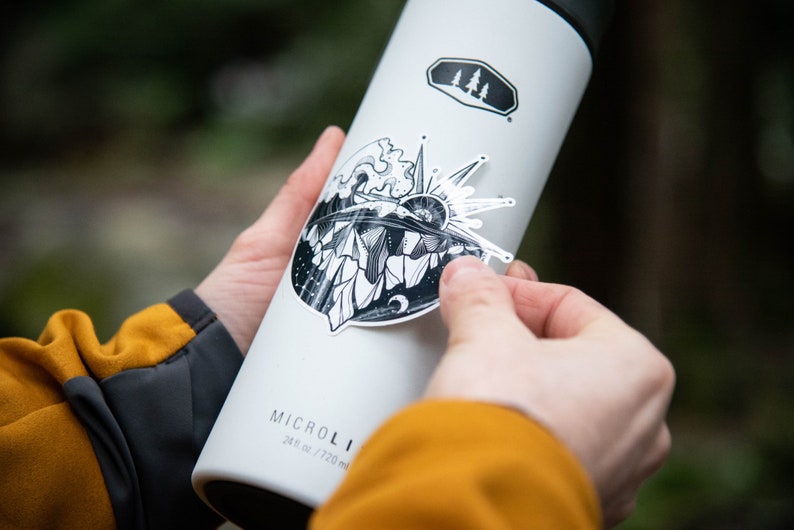 May include: A white stainless steel water bottle with a black and white sticker of a mountain scene with a sun and waves. The bottle has the text "MICRO" and "24 oz / 720 ml" printed on it.